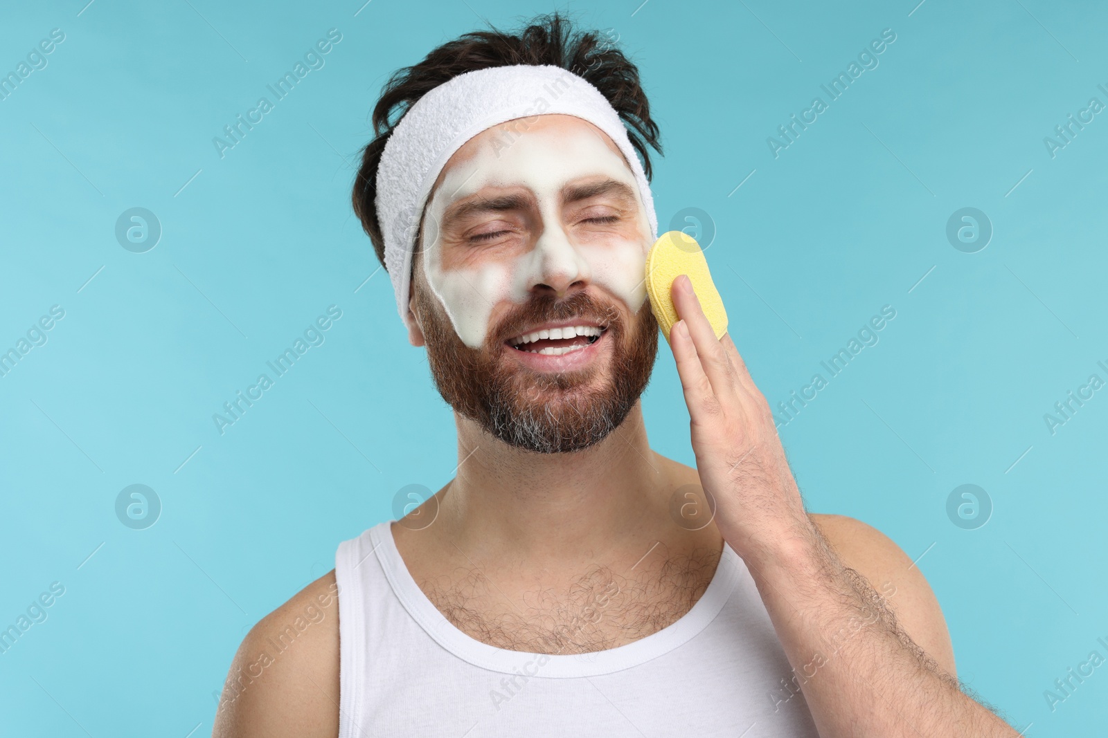 Photo of Man with headband washing his face using sponge on light blue background