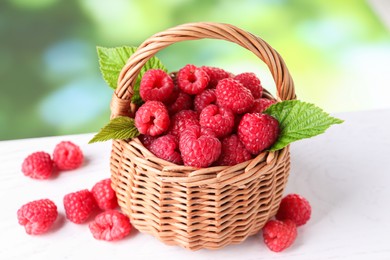 Photo of Wicker basket with tasty ripe raspberries and leaves on white table against blurred green background