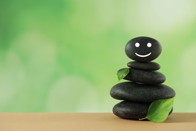 Photo of Stack of stones with drawn happy face and green leaves on table against blurred background, space for text. Zen concept