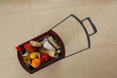 Photo of Shopping basket full of different products on beige tile floor, top view