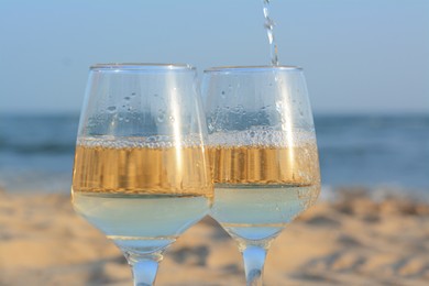 Photo of Pouring white wine into glasses on sandy seashore, closeup