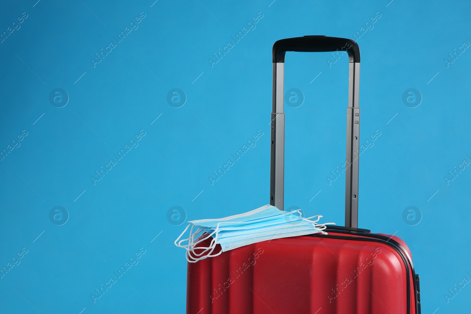 Photo of Stylish red suitcase and protective masks on light blue background, space for text. Travelling during coronavirus pandemic