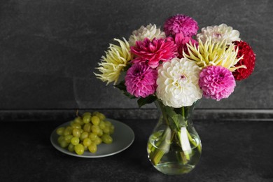 Photo of Bouquet of beautiful Dahlia flowers in vase and plate with grapes on black table near grey wall