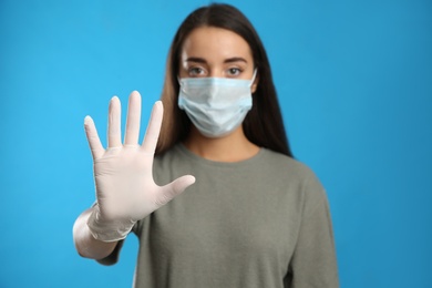 Woman in protective face mask and medical gloves showing stop gesture against blue background, focus on hand