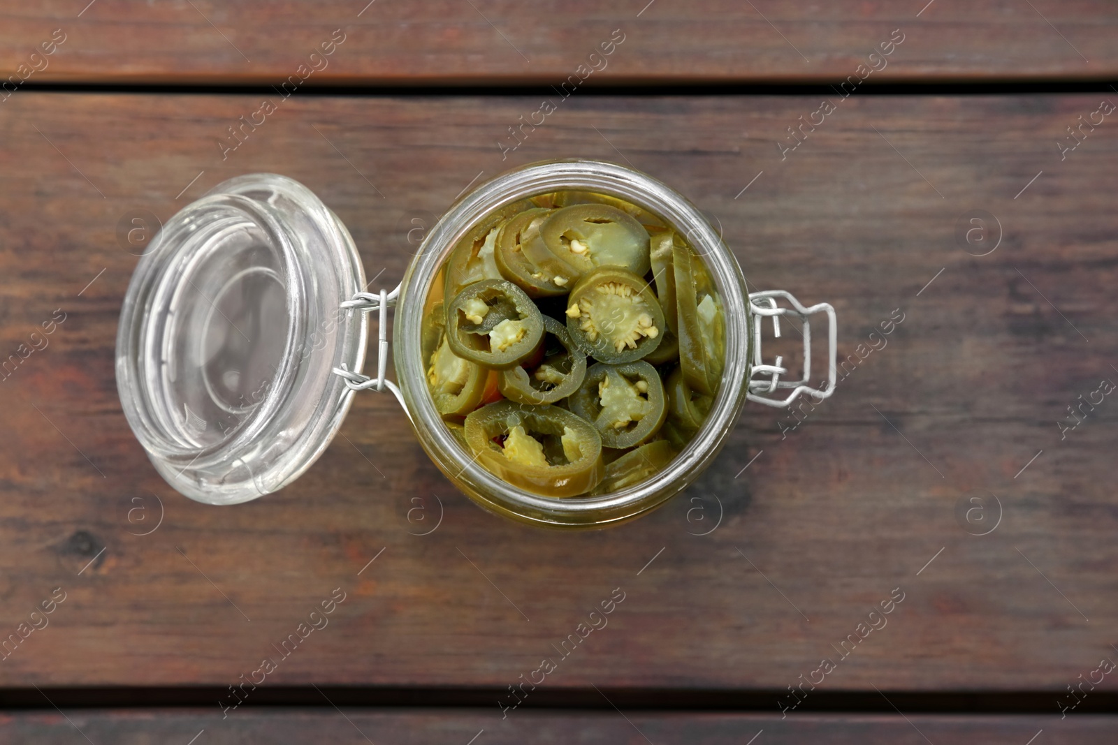 Photo of Glass jar of pickled green jalapeno peppers on wooden table, top view