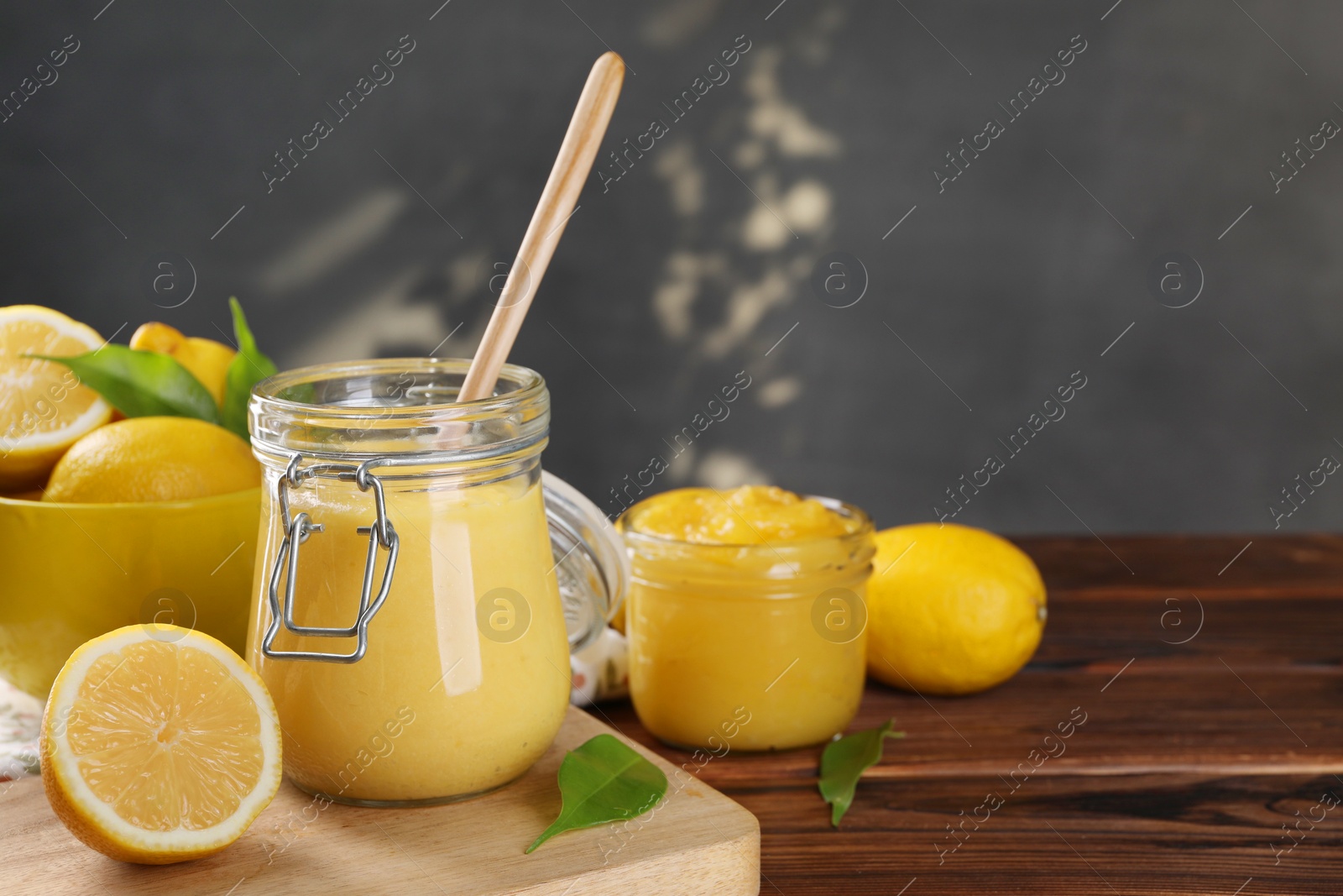 Photo of Delicious lemon curd in glass jars, spoon, fresh citrus fruits and green leaves on wooden table