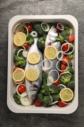 Raw fish with vegetables and lemon in baking dish on grey table, top view