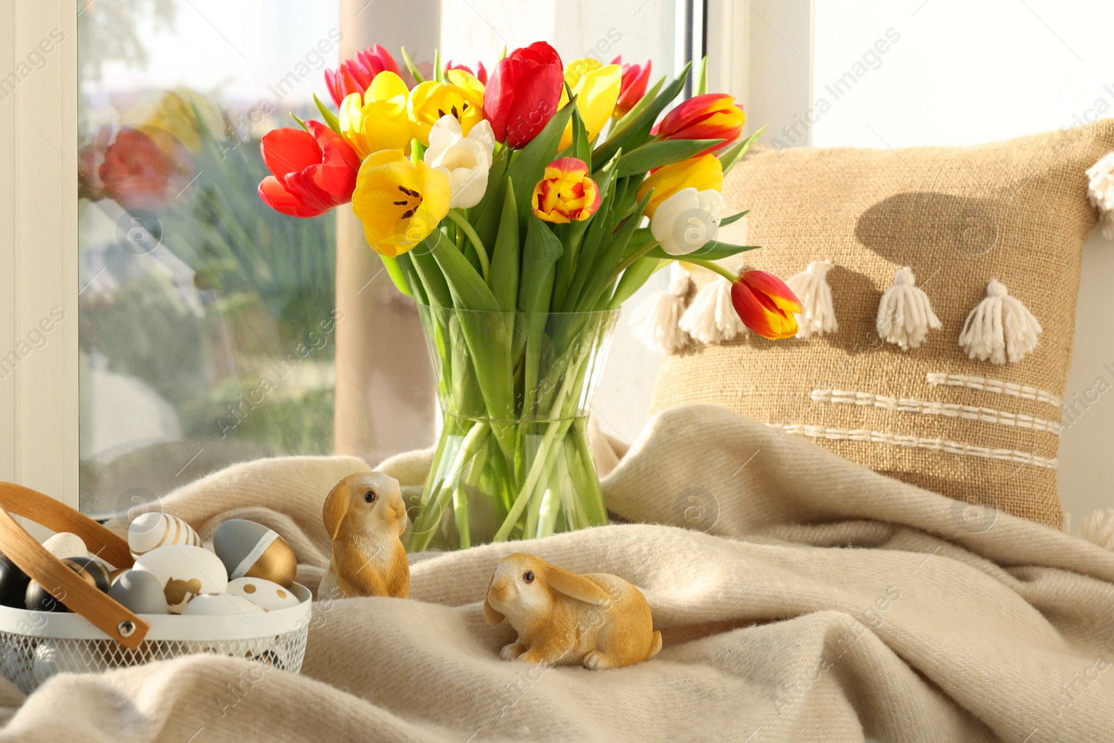 Photo of Easter decorations. Beautiful tulips, basket of painted eggs, bunny figures, pillow and plaid on window sill