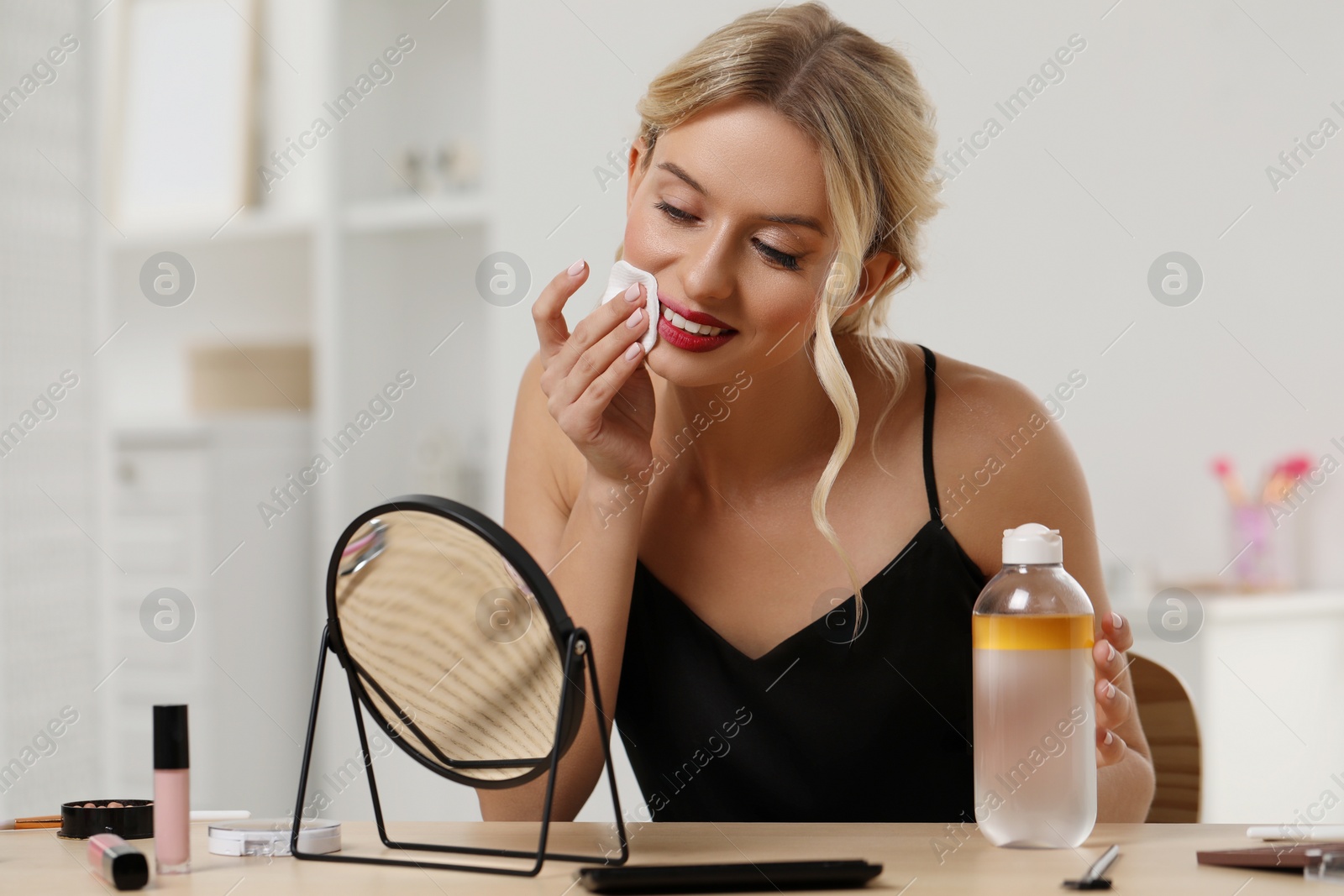 Photo of Smiling woman removing makeup with cotton pad in front of mirror at table indoors
