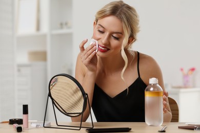 Photo of Smiling woman removing makeup with cotton pad in front of mirror at table indoors