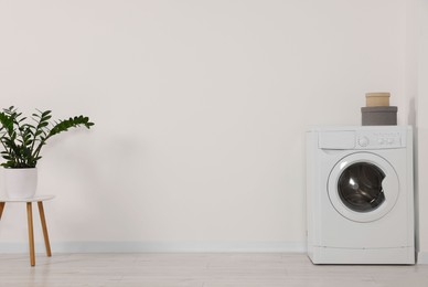 Photo of Modern washing machine and potted plant on stool against white wall indoors