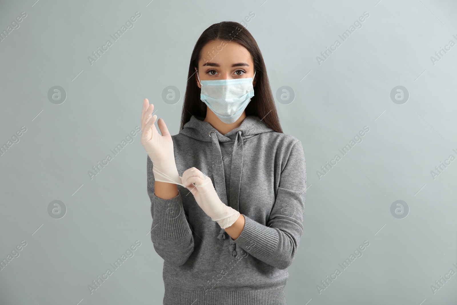 Photo of Woman in protective face mask putting on medical gloves against grey background