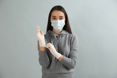 Woman in protective face mask putting on medical gloves against grey background