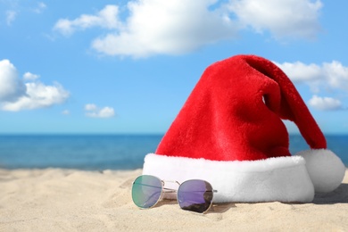 Santa's hat and sunglasses on sandy beach, space for text. Christmas vacation
