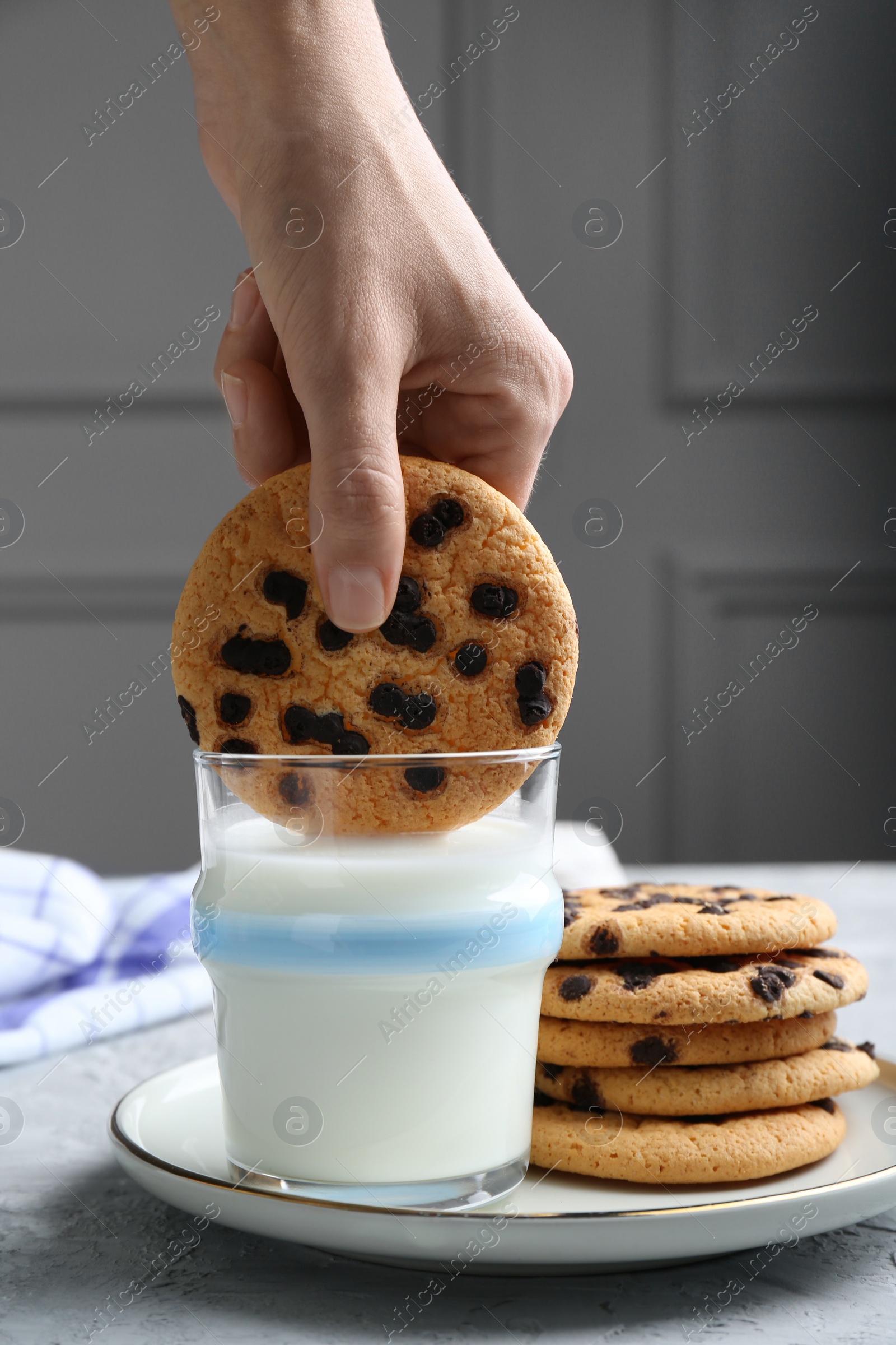 Photo of Woman dipping delicious chocolate chip cookie into glass of milk at grey textured table, closeup