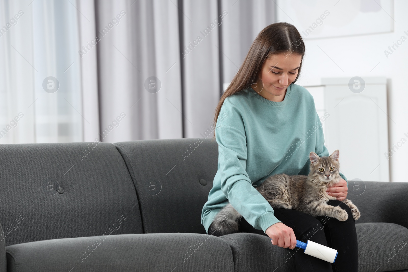 Photo of Pet shedding. Woman with lint roller removing cat`s hair from trousers on sofa at home, space for text