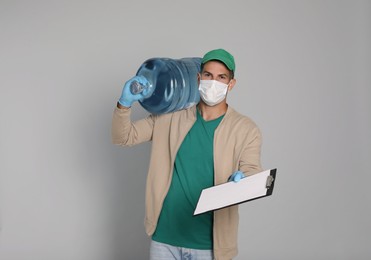 Photo of Courier in medical mask with bottle for water cooler and clipboard on light grey background. Delivery during coronavirus quarantine