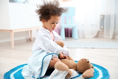 Photo of Cute African American child imagining herself as doctor while playing with stethoscope and toy bunny at home