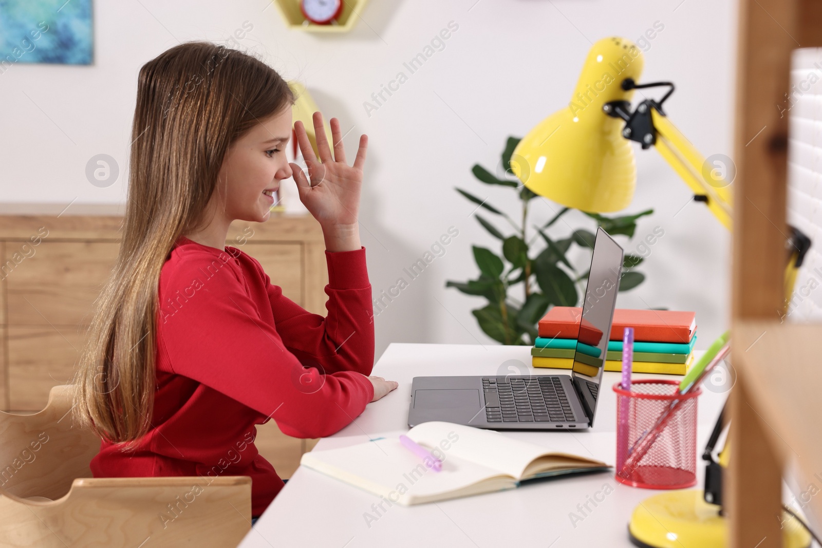 Photo of E-learning. Cute girl raising her hand to answer during online lesson at table indoors