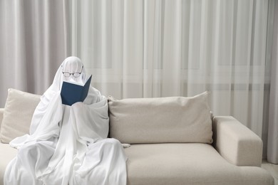 Photo of Creepy ghost. Person covered with white sheet reading book on sofa at home, space for text