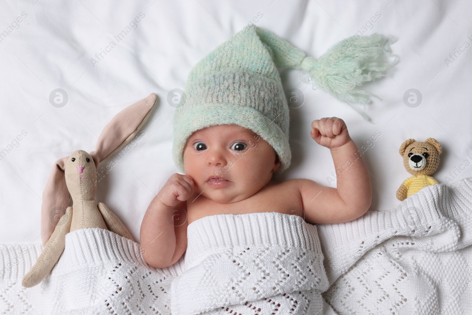 Photo of Cute little baby with toys lying under knitted plaid in bed, top view