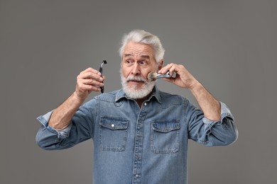 Senior man with mustache holding razor and brush on grey background