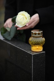 Woman holding white rose near black granite tombstone with candle outdoors, closeup. Funeral ceremony