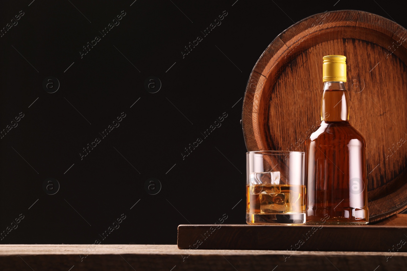 Photo of Whiskey with ice cubes in glass, bottle and barrel on wooden table against black background, space for text