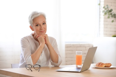 Photo of Mature woman with laptop sitting at table in kitchen. Smart aging
