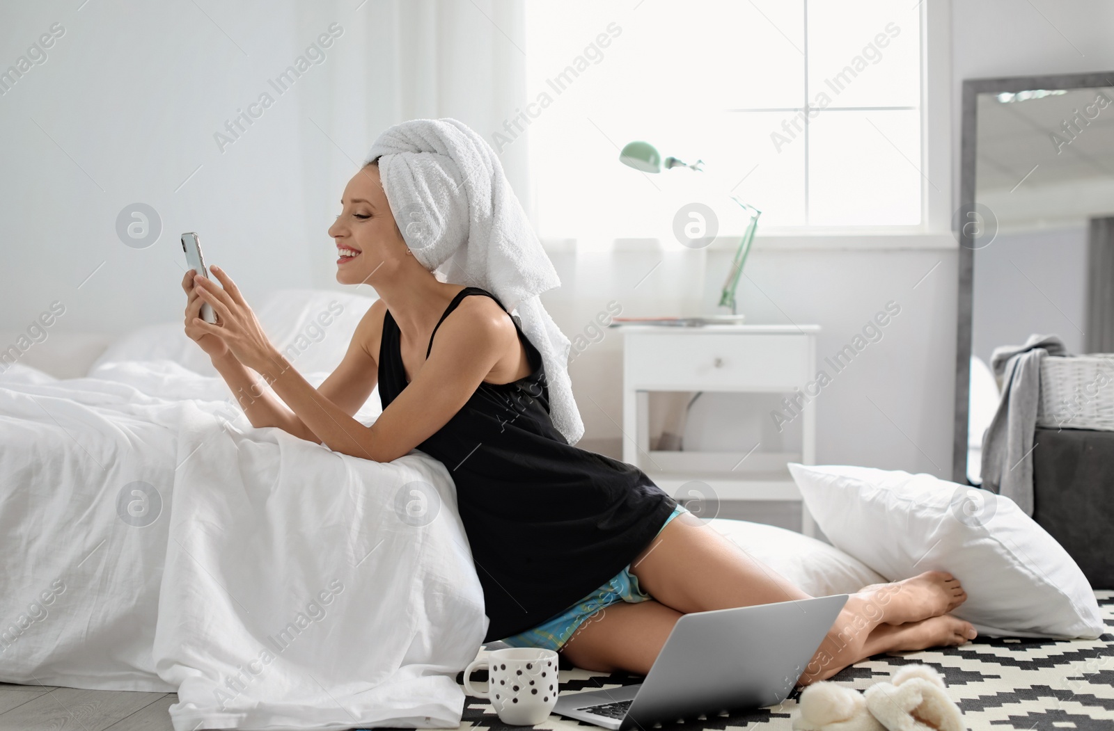 Photo of Young happy woman with mobile phone sitting on floor near bed at home. Lazy morning