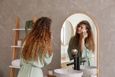 Photo of Beautiful woman cleaning her face with cotton disk near mirror in bathroom