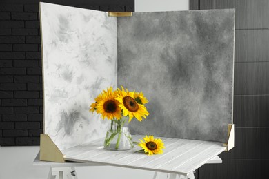 Photo of Glass vase with beautiful sunflowers and double-sided backdrops on table in photo studio