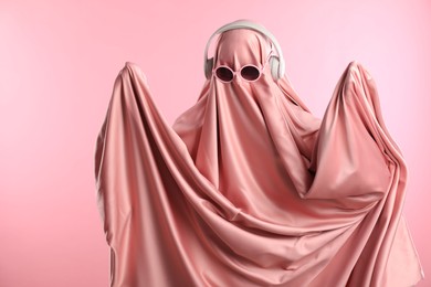 Photo of Glamorous ghost. Woman in sheet with sunglasses and headphones on pink background