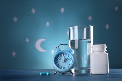 Photo of Alarm clock and soporific pills near glass of water on table against blue wall decorated with stars and crescent, space for text. Insomnia treatment