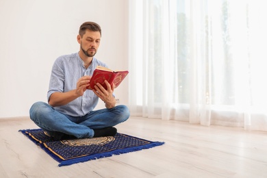 Photo of Muslim man reading Koran on prayer rug indoors. Space for text
