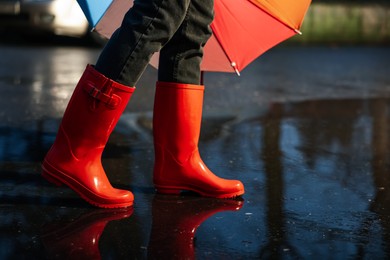Photo of Woman with umbrella and red rubber boots walking in puddle, closeup. Rainy weather