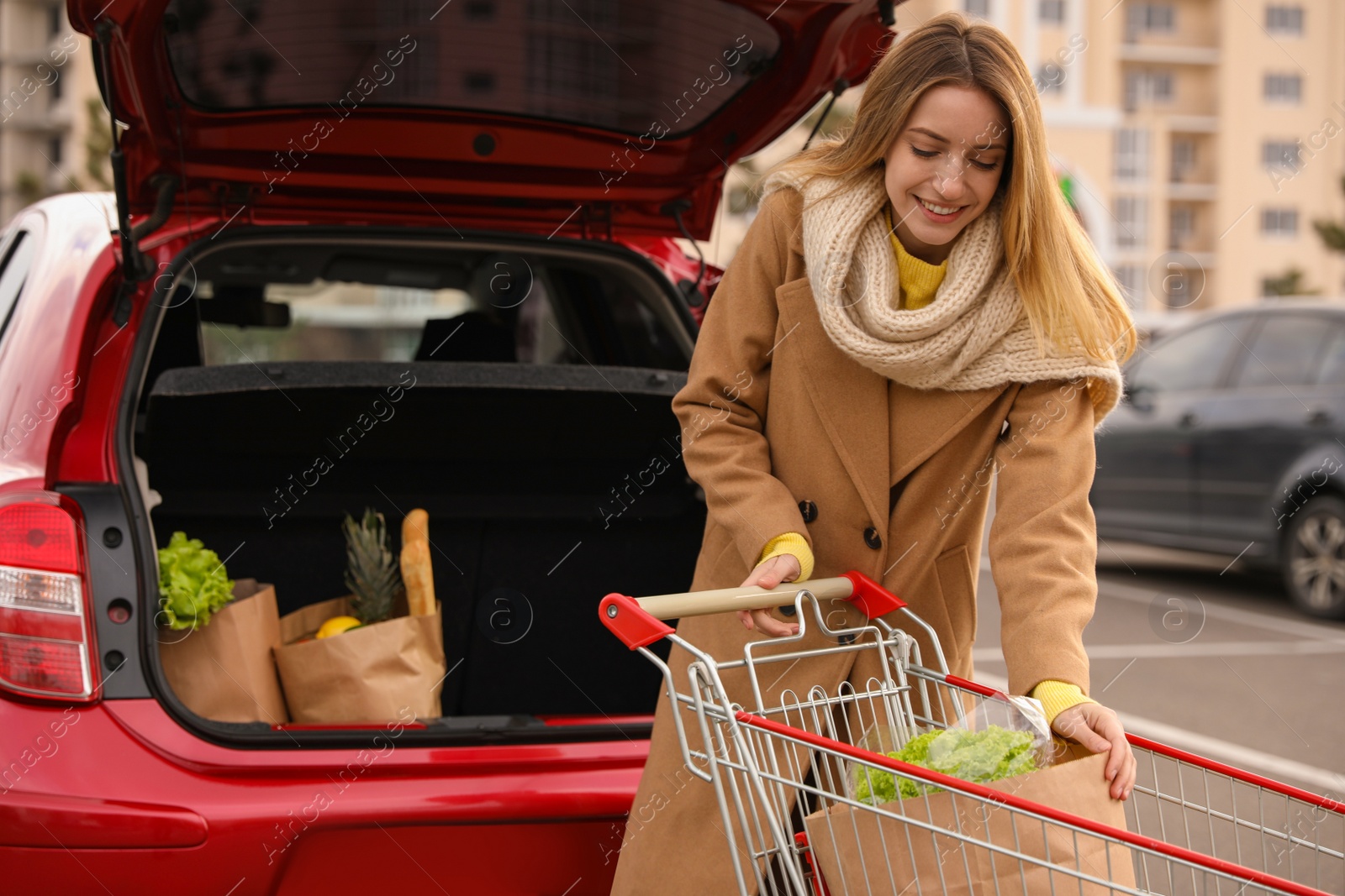 Photo of Young woman putting shopping bags full of groceries into her car outdoors