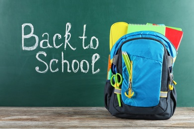 Photo of Backpack with stationery on table against blackboard with written words BACK TO SCHOOL