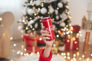 Photo of MYKOLAIV, UKRAINE - January 01, 2021: Woman with can of Coca-Cola against blurred Christmas tree at home, closeup