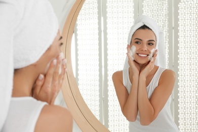 Photo of Happy young woman applying cleansing foam onto face near mirror in bathroom