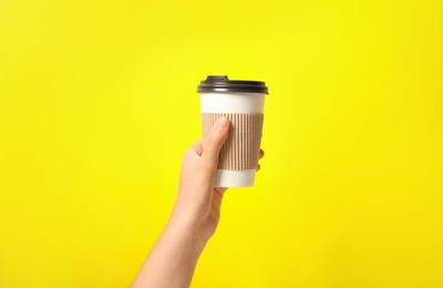 Photo of Woman holding takeaway paper coffee cup with cardboard sleeve on yellow background, closeup