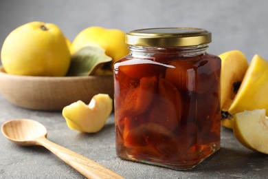 Photo of Tasty homemade quince jam in jar, spoon and fruits on grey textured table, closeup