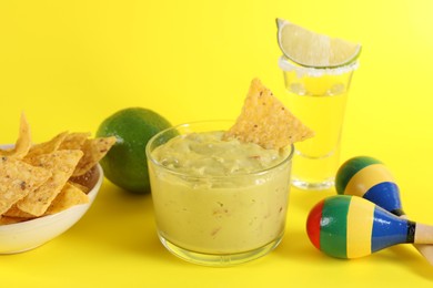 Photo of Delicious guacamole with nachos chips, maracas, tequila and lime on yellow background,