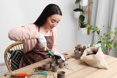 Photo of Young woman adding soil into peat pots at wooden table indoors. Growing vegetable seeds