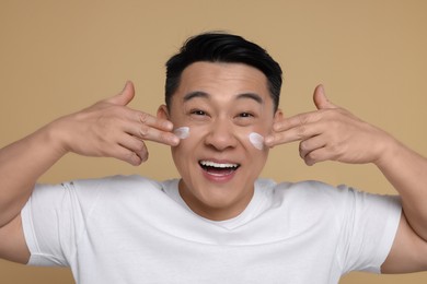 Photo of Emotional man applying cream onto his face on light brown background