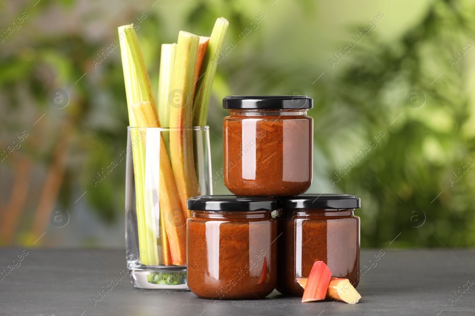 Photo of Jars of tasty rhubarb jam and stalks on grey table, closeup