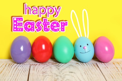 Image of Happy Easter. Blue egg with drawn face and ears as bunny among others on white wooden table against yellow background