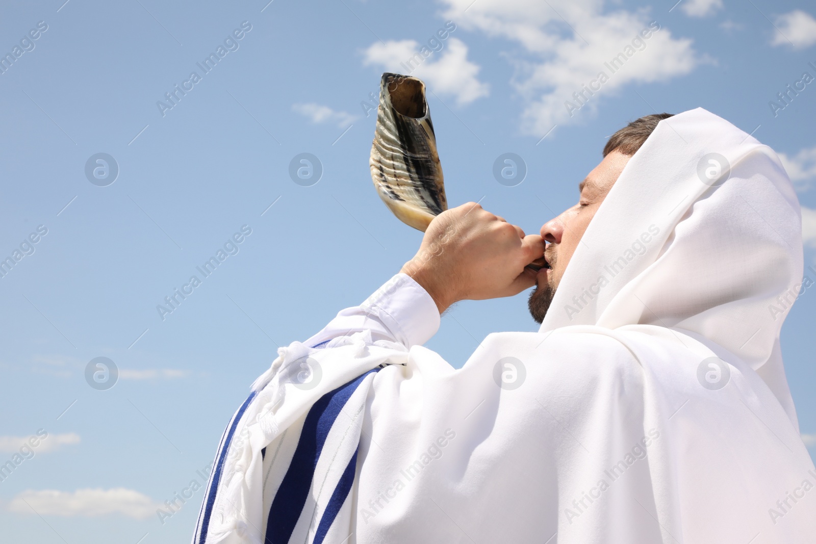 Photo of Jewish man in tallit blowing shofar outdoors. Rosh Hashanah celebration