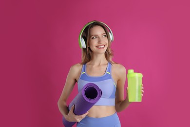 Photo of Beautiful woman with headphones, yoga mat and shaker on pink background