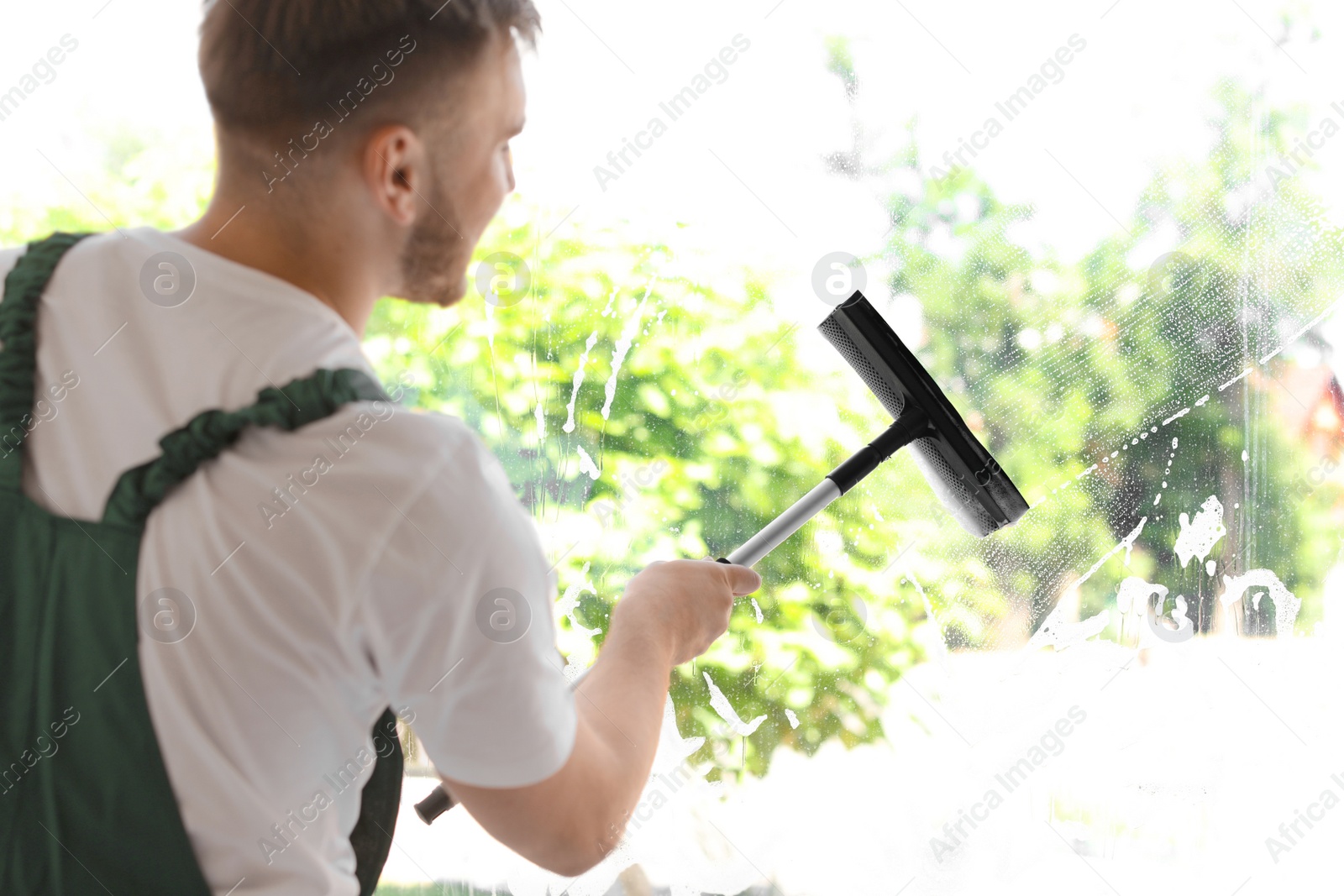 Photo of Male cleaner wiping window glass with squeegee indoors